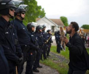 Un manifestante sostiene una pancarta mientras salta en repetidas ocasiones para que los policías que se encuentran al fondo puedan verlo en la casa del agente Derek Chauvin, quien fue despedido de la policía de Minneapolis, a las afueras de Oakdale, Minnesota. Foto: AP.