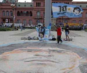 A man waves a flag near a giant portrait of Diego Maradona painted on the ground of historic Plaza de Mayo, near the Government House where fans pay tribute to late football legend Diego Armando Maradona in Buenos Aires, on November 26, 2020. - Diego Maradona's coffin arrived at the presidential palace in Buenos Aires for a period of lying in state, TV reports showed, following the death of the Argentine football legend aged 60 on November 25. Hundreds of people were already lining up to pay their respects to Maradona, who died while recovering from a brain operation, the images from sports channels TyC and ESPN showed. (Photo by ALEJANDRO PAGNI / AFP)