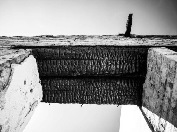 Detalle en escorzo de dos columnas sosteniendo una viga de madera quemada, el ángulo remite a fuerza y resistencia.
