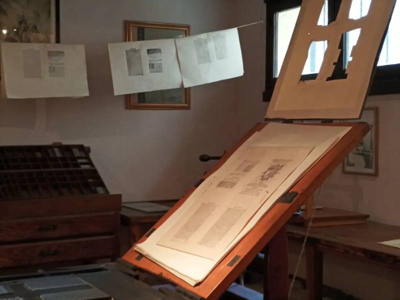 Una hoja impresa se revisa sobre un atril en el taller, donde aún se muestran las técnicas tipográficas del siglo de oro. Al fondo, páginas colgadas se secan al aire, como se hacía en tiempos de Cervantes.