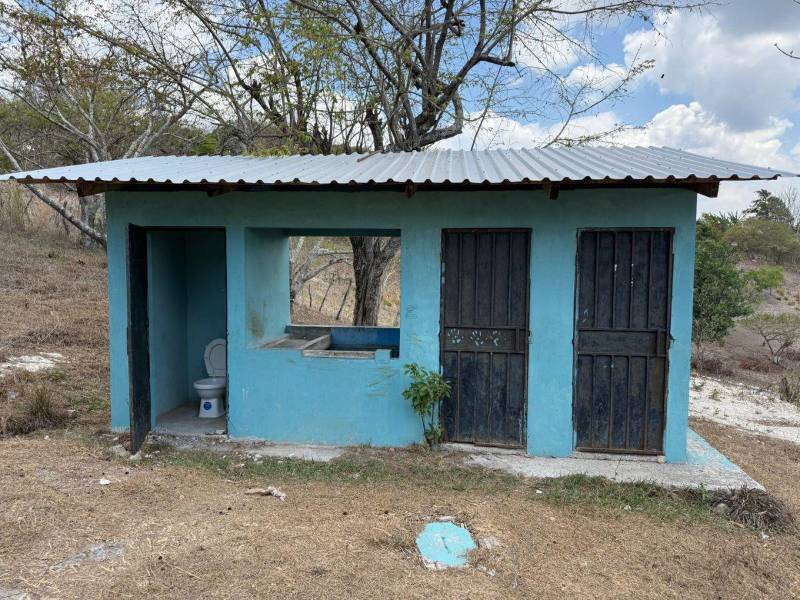 Módulos sanitarios de la escuela Dr. Adalberto Abadie, ubicados a varios metros de las aulas, con puertas metálicas desgastadas y una pila al centro.