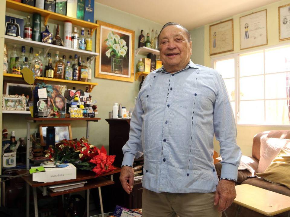 El doctor Augusto Aguilar posando junto a su guaroteca, un espacio donde guarda todas las bebidas que le han regalado en los lugares en los que ha participado como observador electoral.